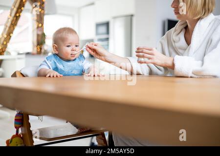 La madre indossa un comodo cucchiaio da bagno che allatta il bambino in poltrona con purea di frutta al tavolo da pranzo a casa. Concetto di introduzione degli alimenti solidi per bambini. Foto Stock