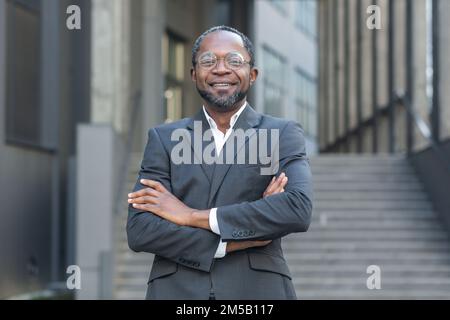 Boss afroamericano riuscito con le braccia incrociate sorridendo e guardando l'uomo maturo della macchina fotografica fuori dell'edificio dell'ufficio. Foto Stock