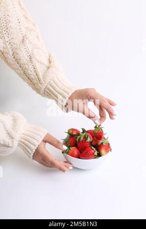 Un colpo verticale di una mano che raccoglie le fragole da una ciotola bianca su una superficie bianca Foto Stock