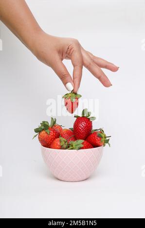 Un colpo verticale di una mano che raccoglie fragole da una ciotola rosa su una superficie bianca Foto Stock