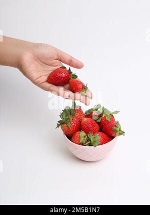 Un colpo verticale di una mano che raccoglie fragole da una ciotola rosa su una superficie bianca Foto Stock