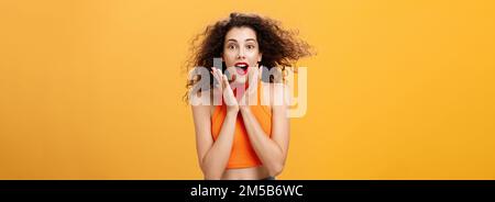 Girata in vita di adornata entusiasta ragazza attraente con acconciatura ricurlato in cima a crop che tiene palme sulle guance e sorridente stupito e. Foto Stock