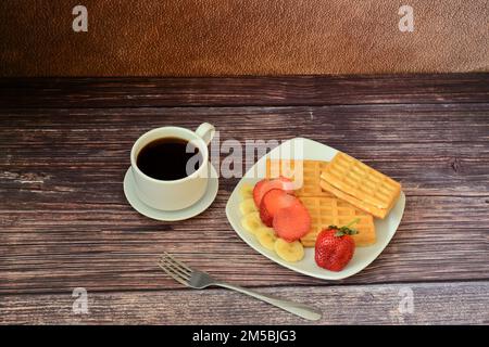 Una tazza di caffè caldo e un piatto di waffle freschi con fragole e banane su un tavolo di legno. Primo piano. Foto Stock