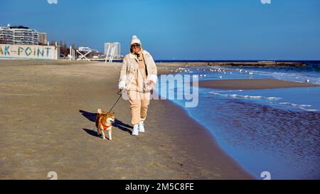 Donna abbastanza matura in una tuta beige camminando lungo la spiaggia con un cane Shiba Inu Foto Stock