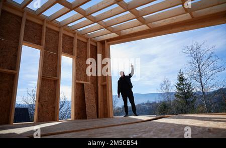 Sviluppatore maschio edificio in legno telaio casa in stile scandinavo barnhouse. Uomo calvo in piedi sul cantiere, che ispeziona la qualità del lavoro nelle giornate di sole con il cielo blu sullo sfondo. Foto Stock