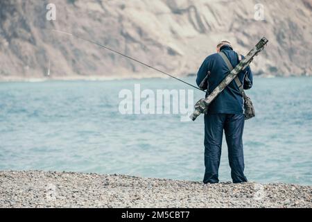 L'uomo hobby pesca sul mare stringe una bobina di lenza di pesce estate. Mare di superficie calmo. Primo piano di un pescatore mani ruota bobina con lenza su Foto Stock