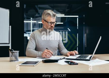 Uomo d'affari serio e pensante all'interno dell'ufficio al lavoro con un computer portatile, un uomo anziano dai capelli grigi che lavora seduto alla scrivania. Foto Stock