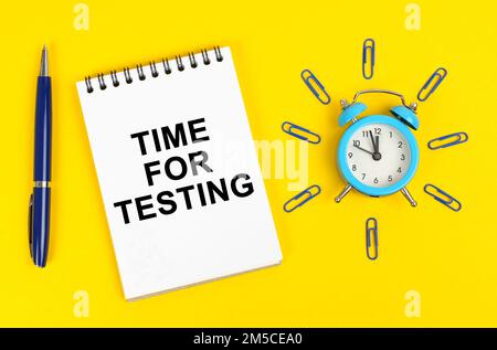 Concetto di business e di istruzione. Su una superficie gialla c'è un orologio di allarme, una penna e un blocco note con l'iscrizione - tempo per il test Foto Stock
