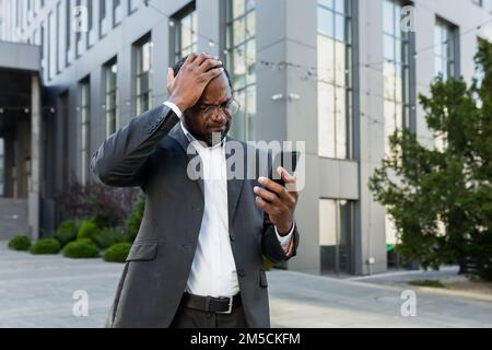 Il capo africano americano maturo che legge le notizie difettose sul telefono, uomo d'affari nel vestito di affari fuori dell'edificio dell'ufficio usando lo smartphone sconvolto e deluso dalla notifica. Foto Stock