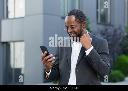 African american businessman boss al di fuori di un moderno edificio di uffici utilizzando il telefono, senior man celebrare la vittoria successo leggere buone notizie online da smartphone, tenendo mano in su gesto di trionfo . Foto Stock