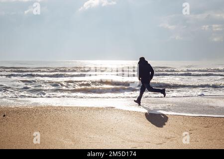 Un uomo che corre lungo la spiaggia di sabbia in una giornata invernale di sole. Buon fine settimana. Foto Stock