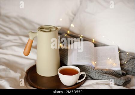 Teiera con tazza di tè e libro di carta aperto su vassoio di legno sopra luce di luce nel letto sulle lenzuola bianche del piumino all'interno. Buongiorno. Ora della colazione. Foto Stock