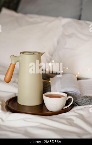 Teiera con tazza di tè e libro di carta aperto su vassoio di legno sopra luce di luce nel letto sulle lenzuola bianche del piumino all'interno. Buongiorno. Ora della colazione. Foto Stock