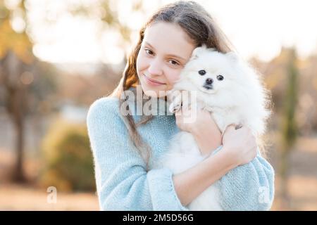 Ragazza giovane sorridente cute 15-16 anni tenendo bianco soffice cane cucciolo spitz insieme su sfondo giallo autunno natura nel parco. Amicizia. Foto Stock