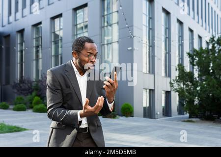 Arrabbiato capo afroamericano litigare e gridare mentre si parla al telefono, uomo anziano in tuta aziendale fuori sede ufficio. Foto Stock