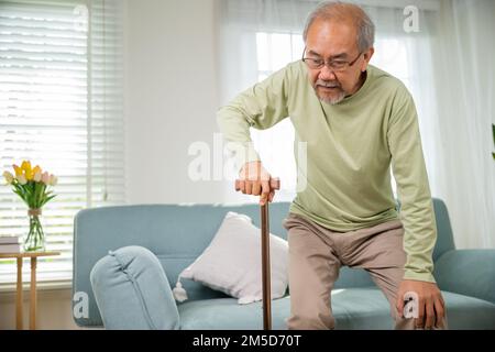 Asian Old Man con occhiali che battono per alzarsi dal divano con bastone da passeggio Foto Stock