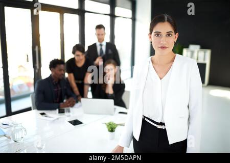 Unisciti a noi per portare il business a un livello superiore. Ritratto di una donna d'affari in piedi in una riunione del consiglio d'amministrazione con i colleghi sullo sfondo. Foto Stock