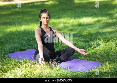 Una giovane donna incinta medita seduta in posizione loto con jnana mudra, pratica yoga su un tappetino nel parco in uno sport nero monopezzo jumpsui Foto Stock