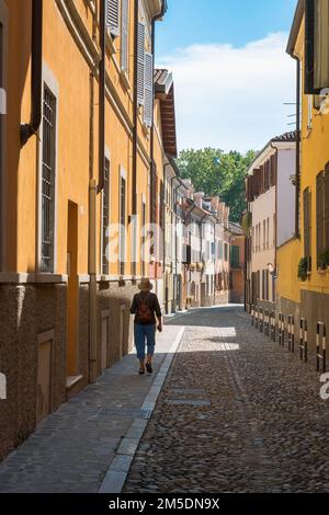 Vacanza donna sola, vista posteriore di una donna di mezza età che cammina da sola in una strada acciottolata nella panoramica zona della città vecchia di Mantova, Italia Foto Stock