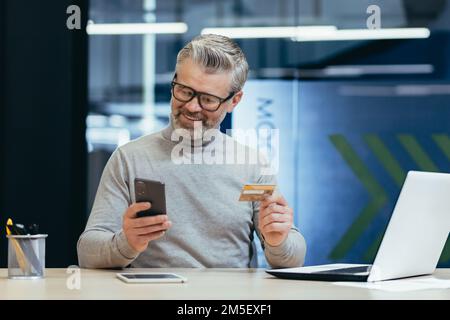 Allegro e sorridente uomo d'affari dai capelli grigi all'interno di un moderno ufficio fa acquisti in un negozio online, l'uomo tiene una carta di credito e uno smartphone nelle sue mani, l'acquirente è scelta soddisfatta. Foto Stock