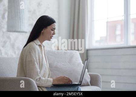 Giovane donna sconvolta ha ricevuto cattive notizie, documenti, lettera di divorzio attraverso la posta online. Si siede triste a casa sul divano, tiene un computer portatile, grida. Foto Stock