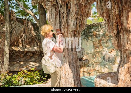 Donna anziana che scatta foto su un telefono cellulare di sculture in un parco naturale. Foto Stock