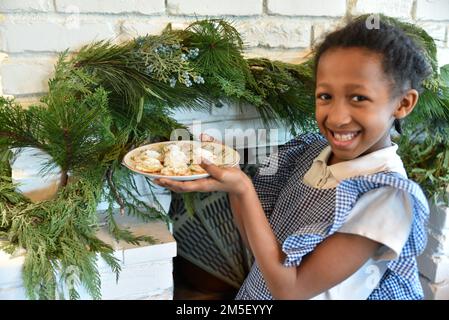 Bambina che tiene un piatto di biscotti appena sfornati fatti in casa il Natale, sorridente grande e felice con verde ghirlanda di Natale drappeggiato nel backgroun Foto Stock