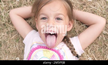 Piccola ragazza sorridente Toothless sorridente che posa sul fieno asciutto dell'erba del prato giallo in mostra la lingua del parco. Estate tempo, natura, Lifestyle Country vita fattoria Foto Stock