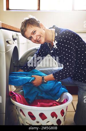 Un lavoro delle madri non finisce mai. Ritratto di una giovane donna allegra che prende il lavaggio da una lavatrice mentre guarda la macchina fotografica a casa. Foto Stock