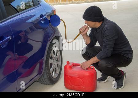 Immagine di un ladro vestito di nero che succhia benzina da un carro armato con un tubo di gomma. Riferimento all'aumento del furto di carburante dovuto a costi eccessivi. Foto Stock