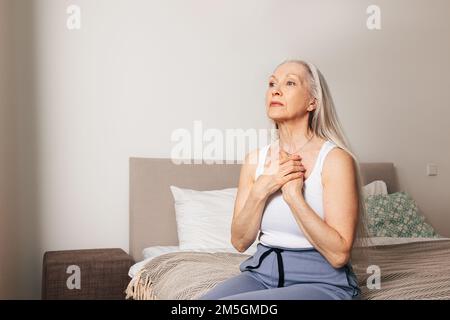 Donna anziana preoccupata seduta su un letto. Donna adulta triste con le mani sul petto. Foto Stock