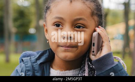 Piccola ragazza etnica che parla sul telefono cellulare all'aperto ritratto divertente bambino multirazziale risposta telefono chiamata piccolo bambino afroamericano figlia Foto Stock