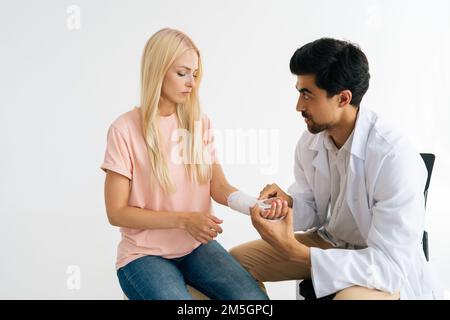 Studio shot di grave medico ortopedico maschile in uniforme esame rotto mano avvolto in cerotto di cerotto di paziente blonde ferito femmina a. Foto Stock