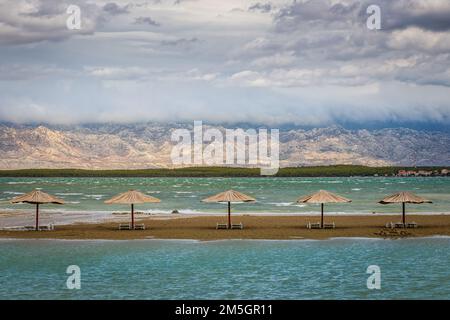 Nin, Croazia - vuota Queen's Beach presso la piccola città mediterranea di Nin alla fine dell'estate con ombrelloni di canna, Velebit Montagne sullo sfondo A. Foto Stock