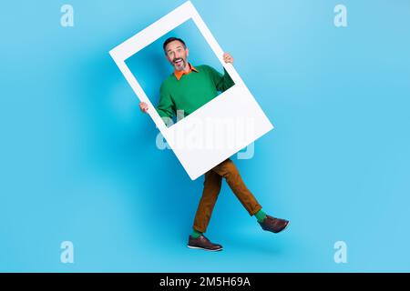 Foto a tutta lunghezza di maturo bel mister camminare posa foto zona cornice finestra indossare abiti verdi alla moda isolato su sfondo di colore blu Foto Stock