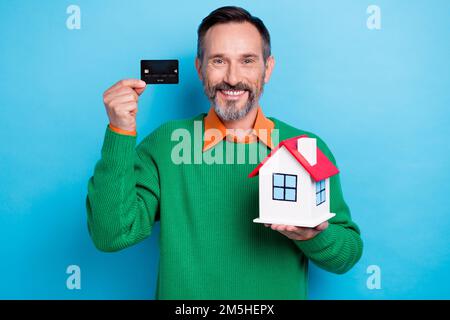 Foto ritratto di maturo bel mister che tiene piccola casa carta di credito vestito elegante verde vestiti isolati su sfondo di colore blu Foto Stock