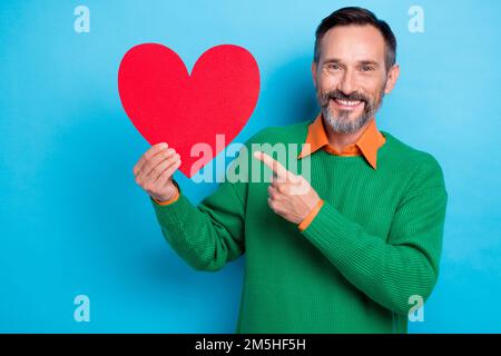 Foto ritratto di maturo bel mister tenere punto rosso cuore forma donazione promo indossare abiti trendy verde isolato su sfondo di colore blu Foto Stock