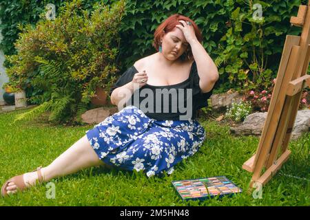 giovane artista caucasica latina, ispanica di etnia argentina, seduta sull'erba nel giardino della sua casa con un cavalletto e gesso colorato Foto Stock