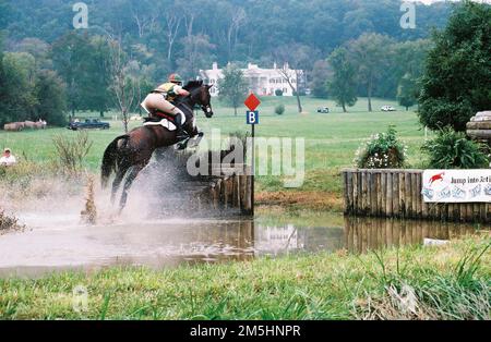 Viaggio attraverso la Hallowed Ground Byway - evento equestre al Morven Park. Con la storica Governor's Mansion in lontananza, un equestre guida il suo cavallo attraverso Poplar Puddle e oltre un ostacolo durante una delle numerose corse di cavalli che si tengono ogni anno presso il Morven Park's International Equestrian Center. Morven Park, Virginia (39,139° N 77,570° W) Foto Stock