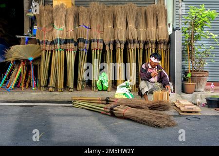 Bangkok, Thailandia, 30 maggio 2022: Un lavoratore lavora su un sentiero di fronte a un negozio che vende scope di cocco e altri strumenti. Foto Stock