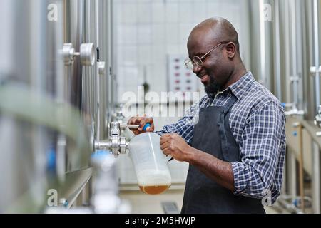 Lavoratore di fabbrica afroamericano che versa la birra prodotta in un contenitore per la degustazione dopo la lavorazione Foto Stock