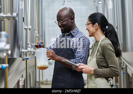 Coppia multietnica di ingegneri che versano birra fresca dal barile per assaggiarla durante il loro lavoro in fabbrica di birra Foto Stock