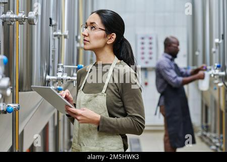 Giovane ingegnere donna che utilizza un tablet digitale per controllare la pressione in barili con birra prodotta Foto Stock