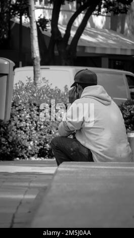 Una scala di grigi verticale di un maschio con cappuccio e felpa con cappuccio, seduto sul marciapiede di una strada a Città del Messico Foto Stock