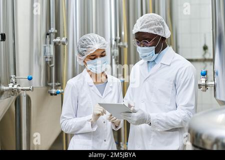 Coppia multietnica di ingegneri in abbigliamento protettivo che lavorano su PC tablet in team mentre si trovano in officina con barili di acciaio in background Foto Stock