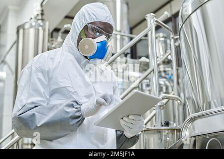 Chimico afroamericano in costume protettivo che lavora online su tablet pc durante il suo lavoro sulla produzione chimica Foto Stock