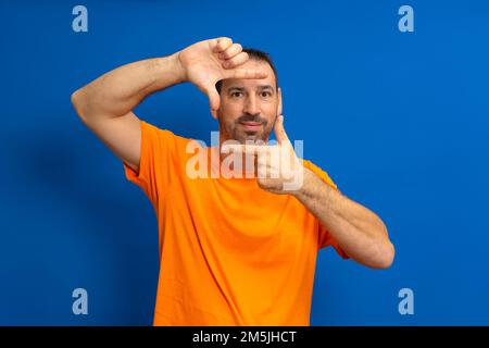 Bel maschio felice bearded gli occhi e guarda attraverso una cornice formata dalle sue mani. Un uomo attraente fa una cornice con le sue dita, isolato Foto Stock