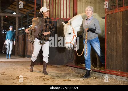 Donna anziana rancher che porta cavallo fuori stalla, mentre donna asiatica in sella Foto Stock