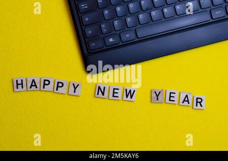 la parola felice anno nuovo stampata su pezzi di legno accanto a una tastiera su sfondo giallo. Concetto IT per l'anno nuovo Foto Stock