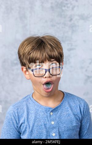 un ragazzino con gli occhiali che fanno un volto sciocco con una lingua blu che sporge e copia spazio Foto Stock